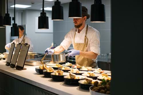 en kokk med hvit jakke og beige forkle heller over saus på varmretter i en kantine