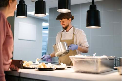 en kokk med hvit jakke, beige forklare og hatt smiler mens han anretter en tallerken med dagens varmrett i en kantine, mens en gjest står i forgrunnen